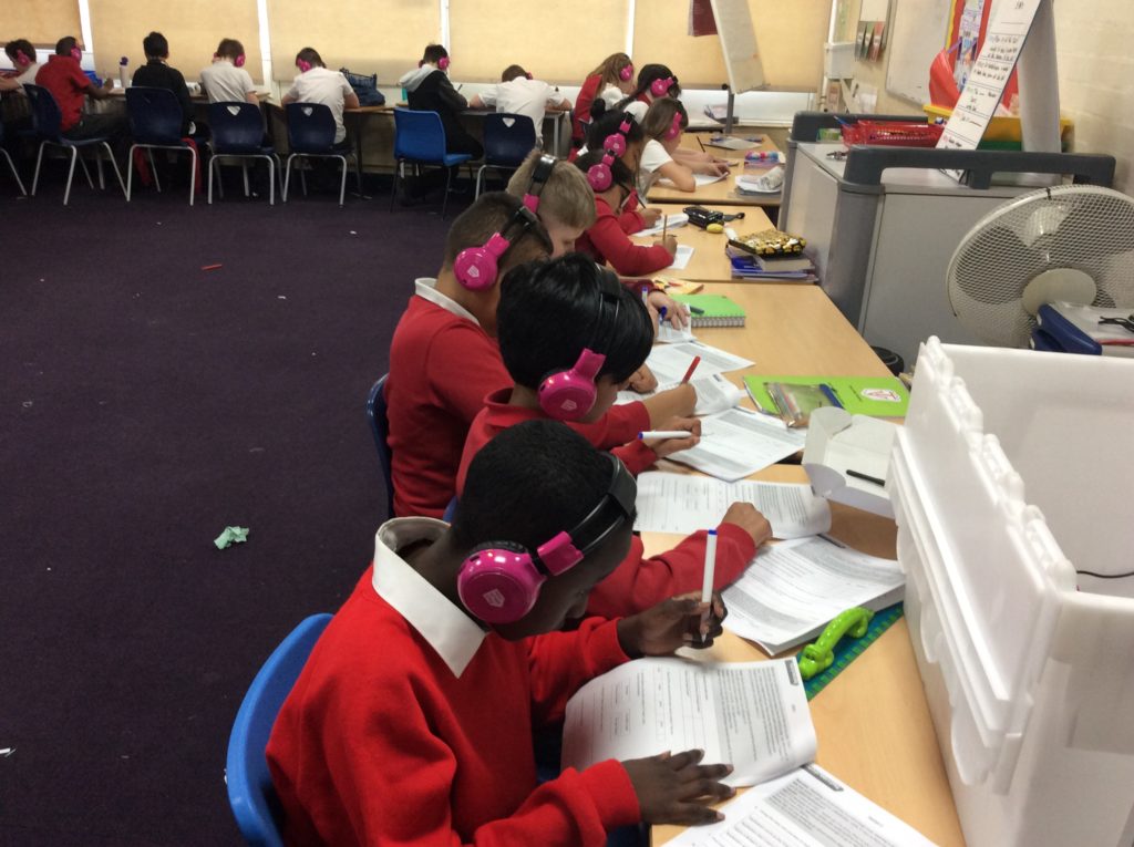 Children wearing now press play headphones sit at desks listening to music whilst doing a written assignment.