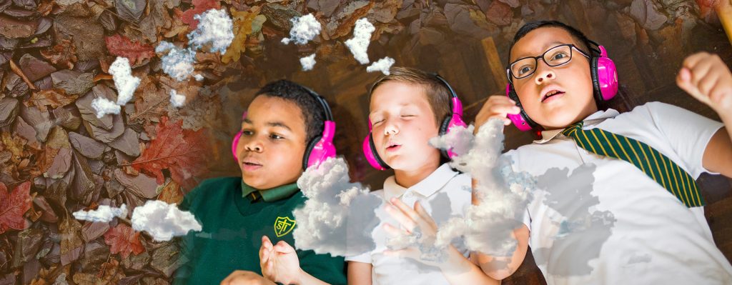 Three boys lie on a wooden school hall floor, which fades into a bed of leaves. They are wearing pink Now Press Play headphones.