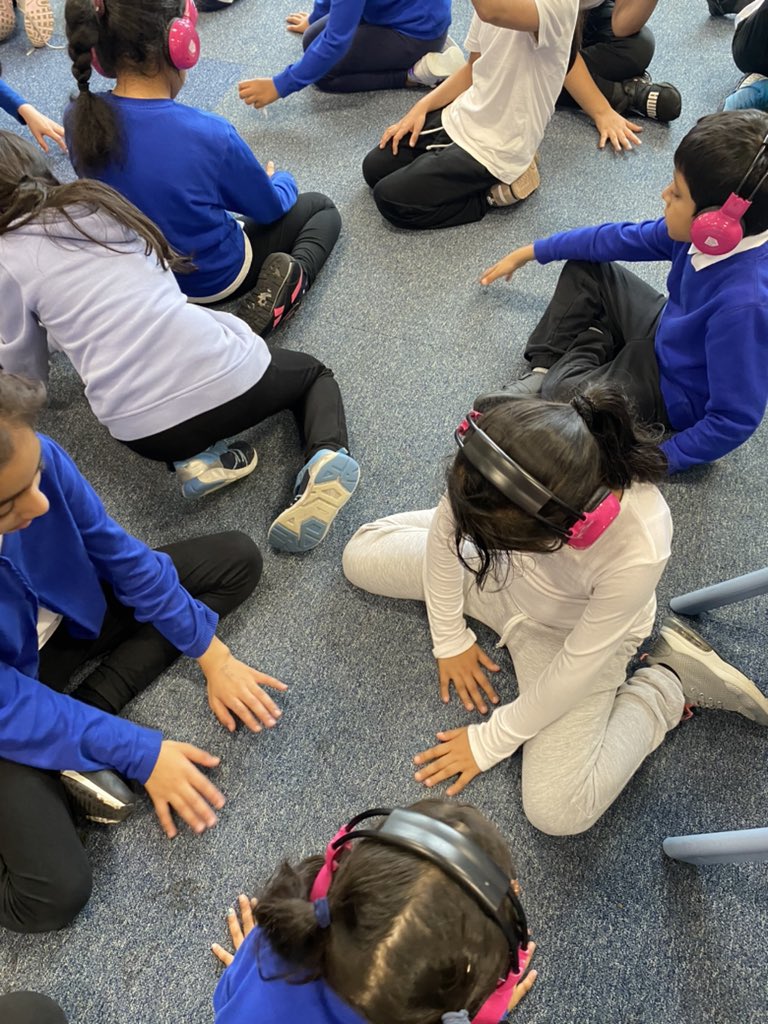A group of children sit on their classroom floor using Now Press Play. They're wearing a blue school uniform and pink headphones.