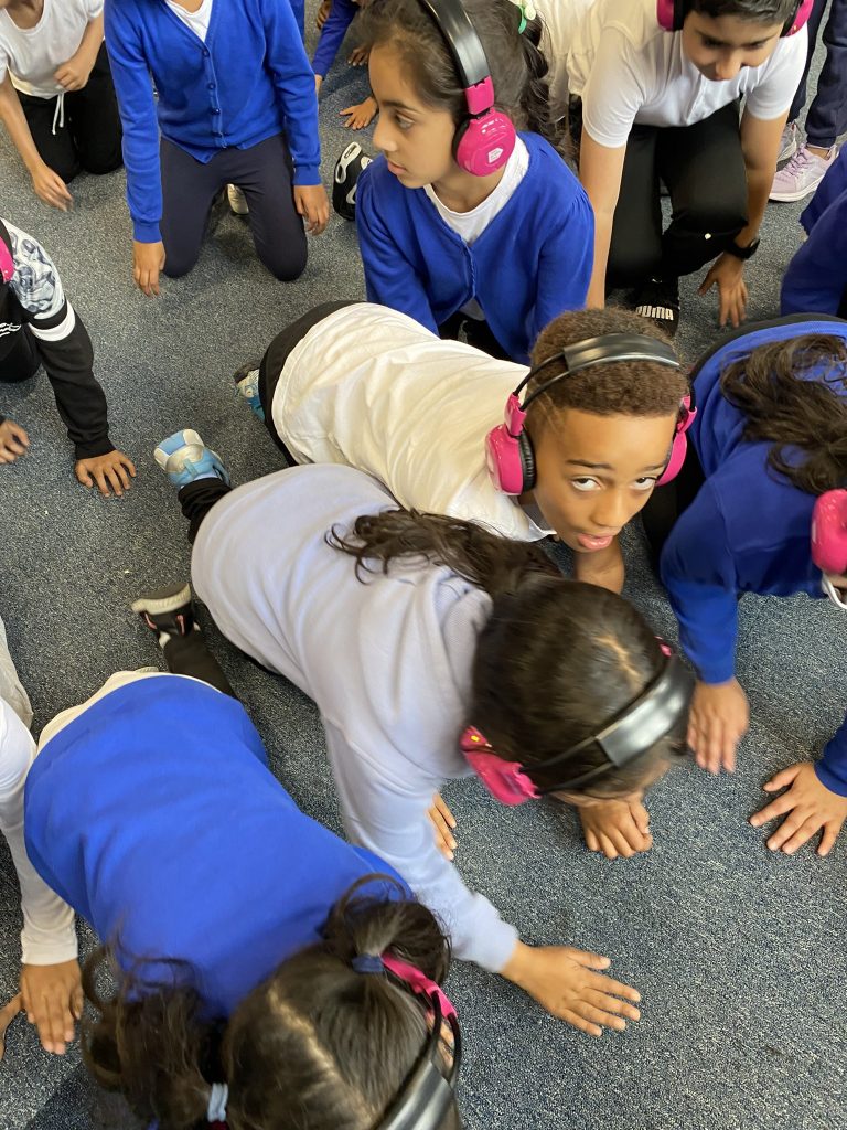 A group of primary school children crawl on a classroom floor listening to Now Press Play. They're wearing a blue school uniform and pink headphones.