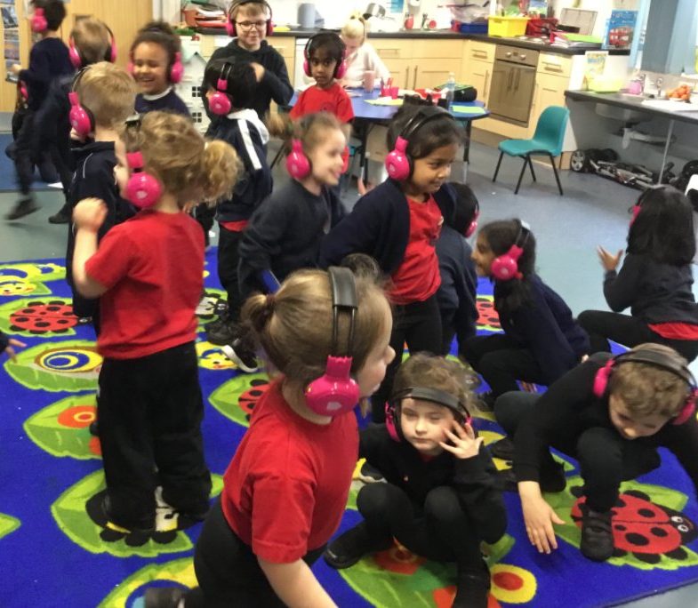 A group of Brookfield Academy students use Now Press Play in their classroom. They're wearing a red uniform and pink headphones. Some of them are crouching and some are standing.