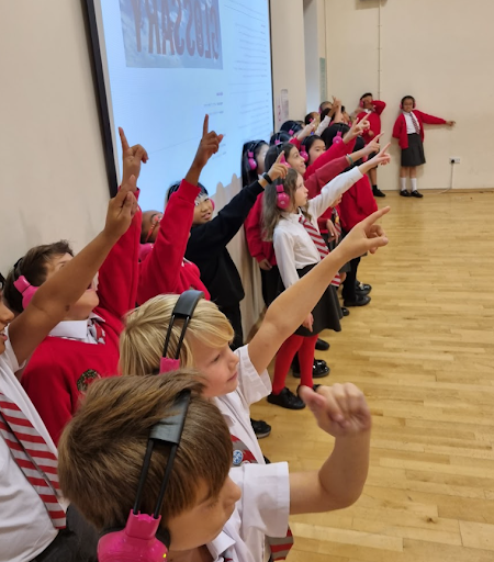 Primary school children from Cheam Academy stand in a line while using Now Press Play. They're wearing a read uniform and pink headphones. Some of them are pointing up.