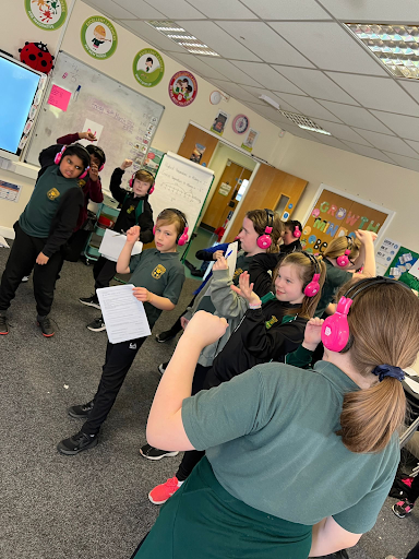 Cheam Farm students stand up in a classroom, using Now Press Play. They're holding worksheets and listening to a Now Press Play experience through pink headphones.