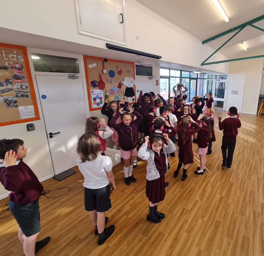 A group of Cheam Fields Academy students walk as a group in a school hall while using Now Press Play. They're wearing a burgundy uniform and pink headphones.
