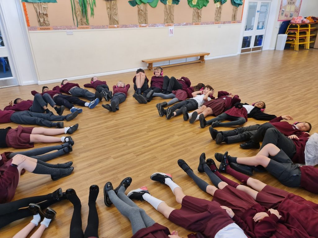 A group of Cheam Fields Academy students lie in a circle in a school hall while using Now Press Play. They're wearing a burgundy uniform and pink headphones.
