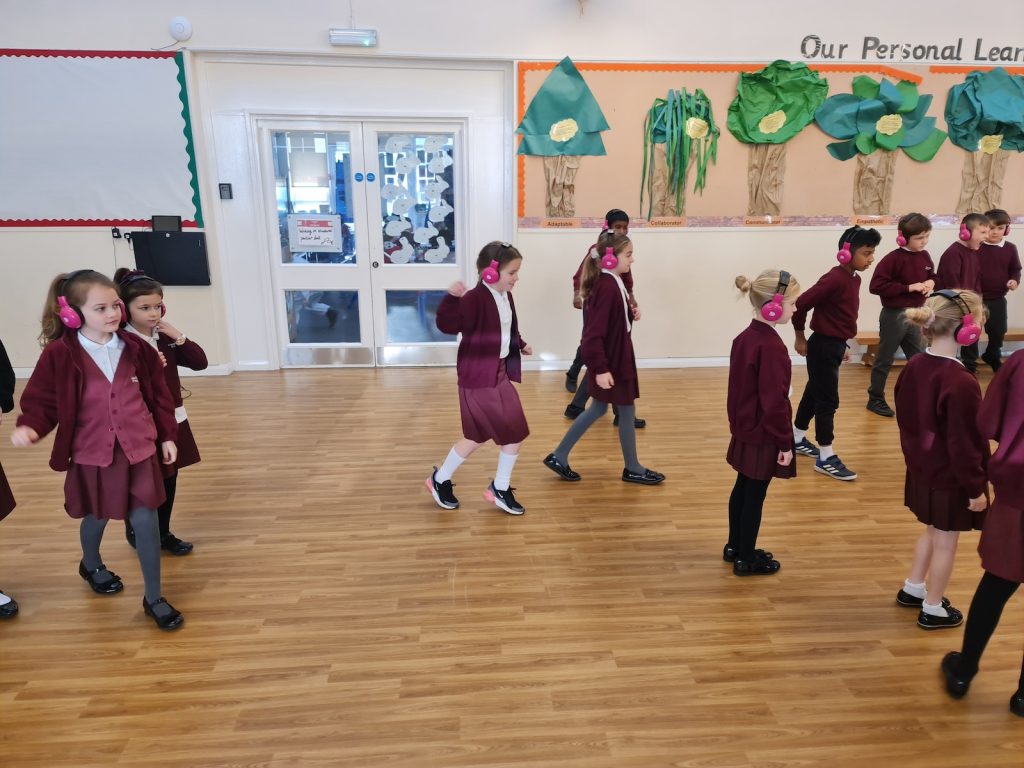 A group of Cheam Fields Academy students walk as a group in a school hall while using Now Press Play. They're wearing a burgundy uniform and pink headphones.
