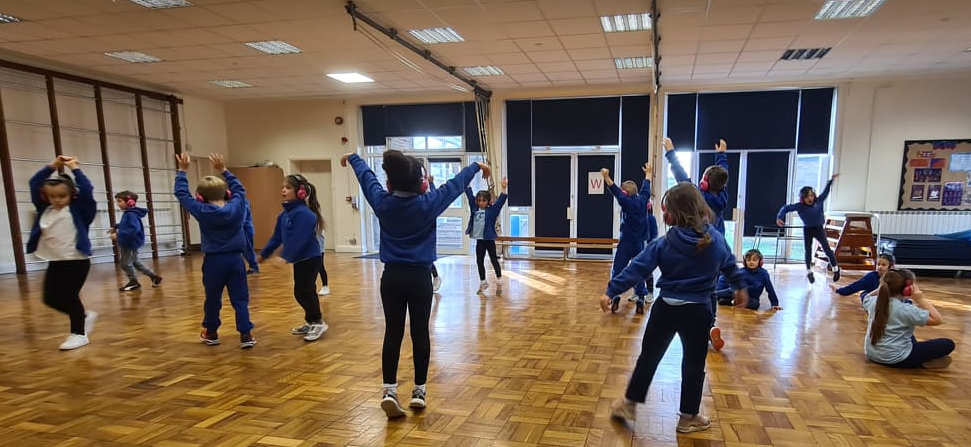 A group of Shawley Community Academy students dance as a group in a school hall while using Now Press Play. They're wearing a burgundy uniform and pink headphones.