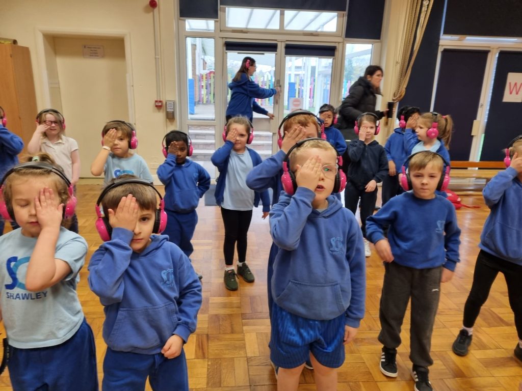 A group of Shawley Community Academy students cover their eyes in a school hall while using Now Press Play. They're wearing a burgundy uniform and pink headphones.