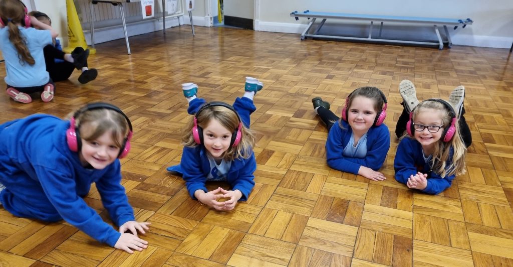 A group of Shawley Community Academy students lie down in a school hall while using Now Press Play. They're wearing a burgundy uniform and pink headphones.