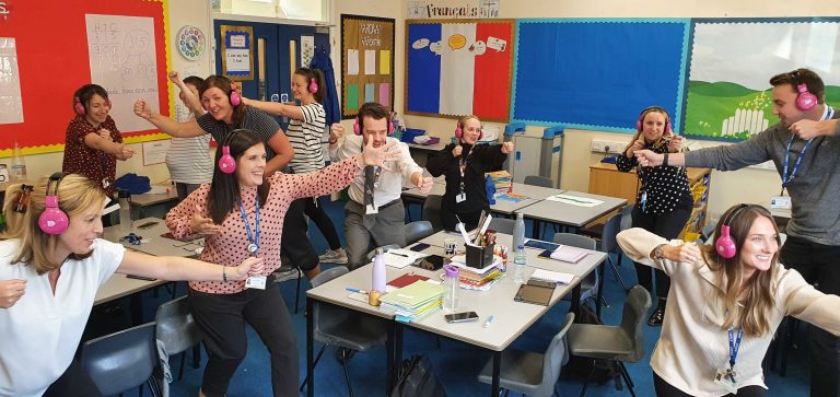 Group of primary school teachers using Now Press Play acting and wearing pink headphones in classroom
