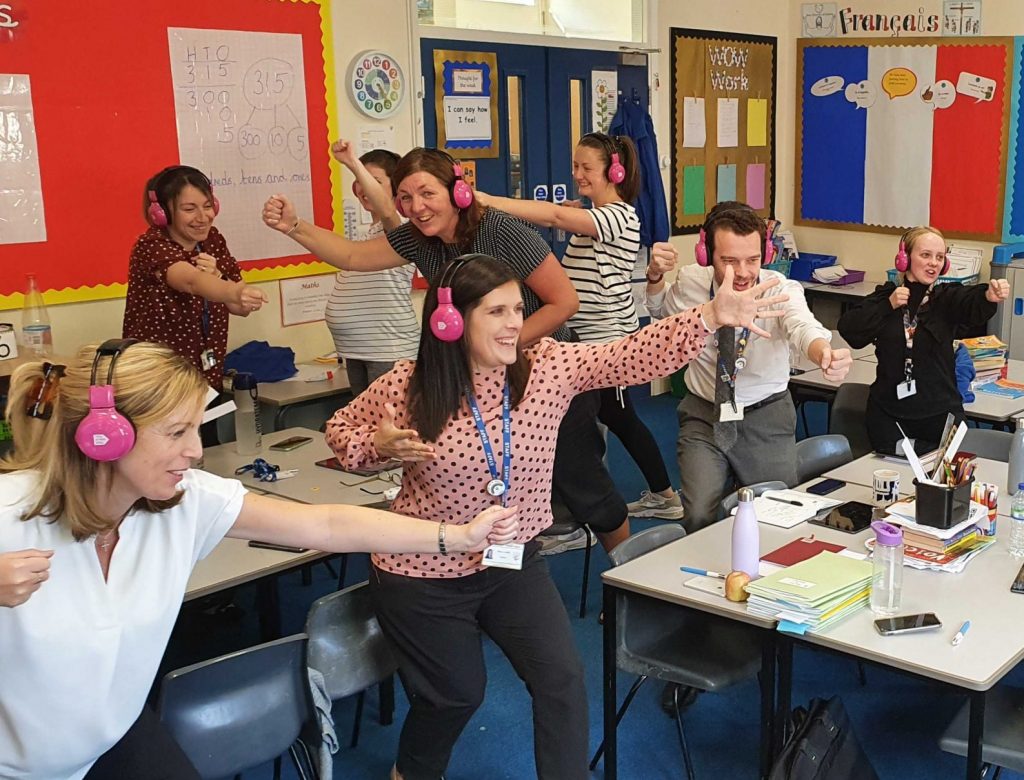 Group of primary school teachers using Now Press Play acting and wearing pink headphones in classroom
