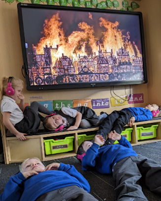 School children use Now Press Play in their classroom. They're lying on the floor pretending to sleep. On the interactive whiteboard is a picture of the Great Fire of London.