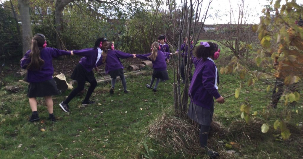 Students from Whitefield Primary School use Now Press Play while outdoors. They're exploring the environment around them together while listening through pink headphones.