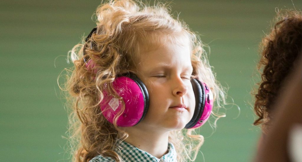 A young school child closing her eyes while using Now Press Play. She's listening through pink headphones.
