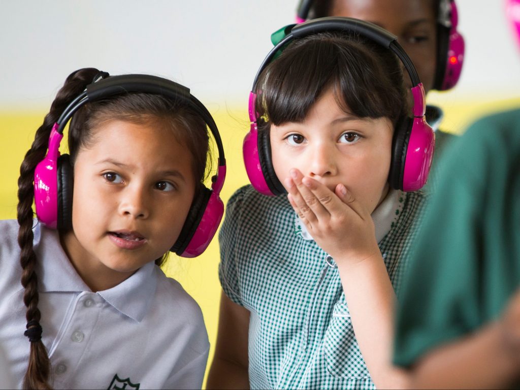 Two young school students use Now Press Play. They look shocked while listening to a lesson through their pink headphones; one of them is covering her mouth in shock.