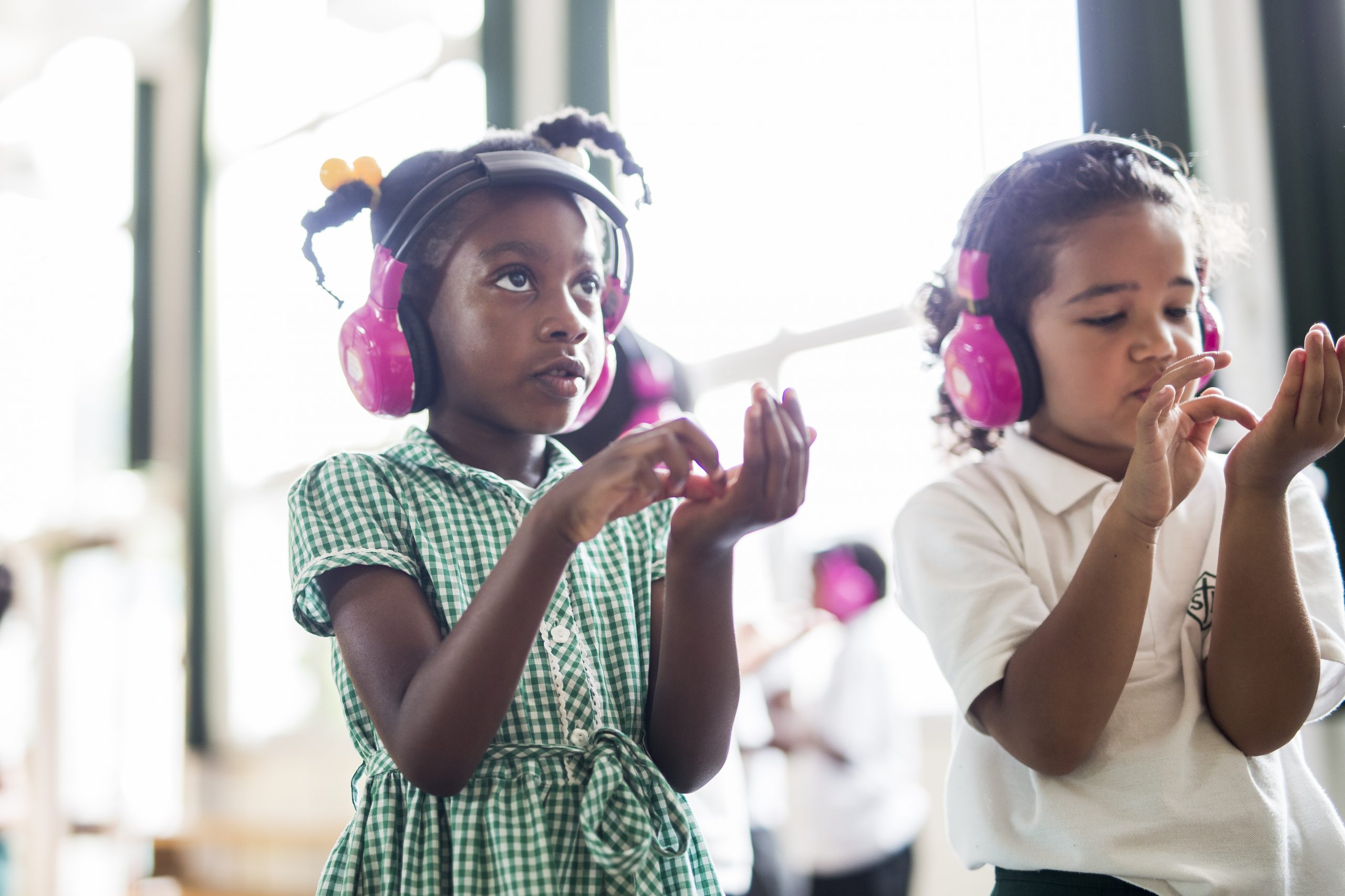 Two young school students use Now Press Play. They're pretending to play the violin while listening to a lesson through their pink headphones.