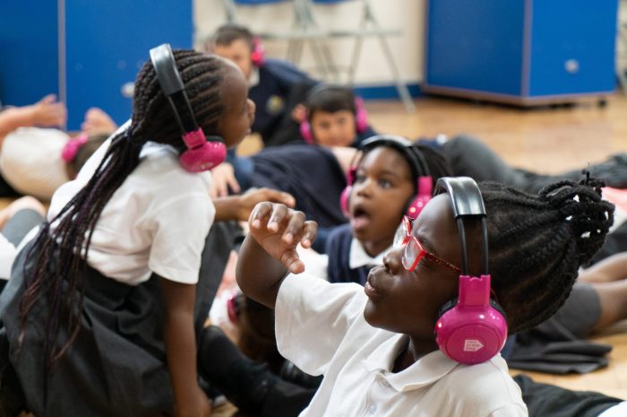 A group of children lie on the floor of a school hall using Now Press Play. They're looking up, and look shocked by what they are visualising through listening through the pink headphones.