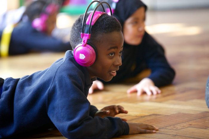 Picture of two children lying on the floor of a school hall using Now Press Play. They are wearing a blue uniform and pink headphones.