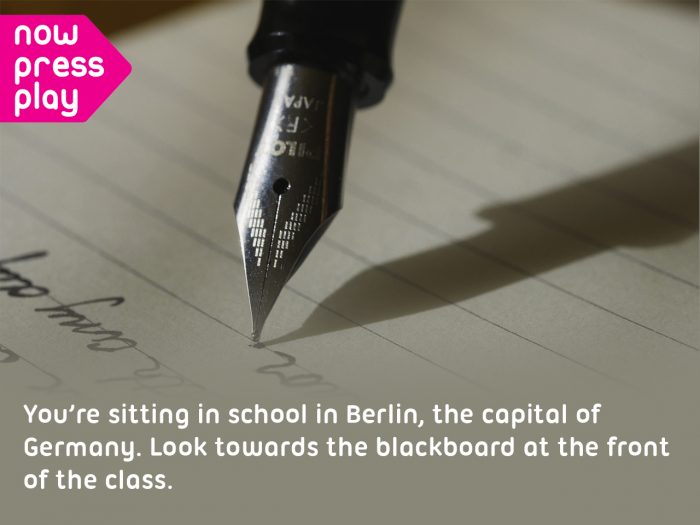 Close up image of a fountain pen writing on paper, from Now Press Play's Relative Clauses Experience. Text reads: You're sitting in school in Berlin, the capital of Germany. Look towards the blackboard at the front of the class.