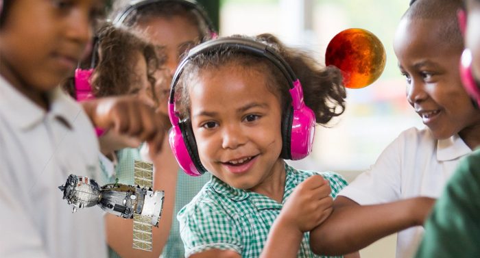 A young girl smiles as she learns science with Now Press Play. She's wearing pink headphones.