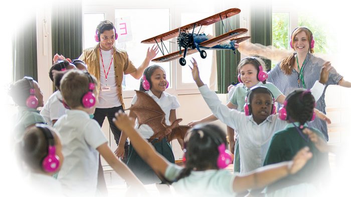Children and teachers in a school hall hold their arms out as they imagine themselves flying while using Now Press Play.