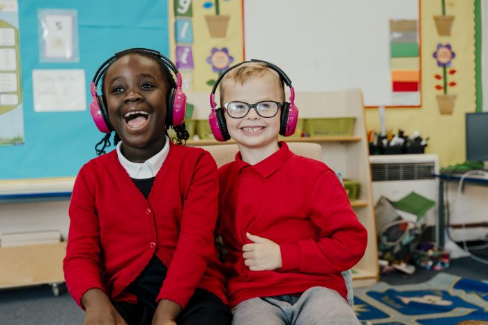 Two primary school children in red uniforms are using Now Press Play in their classroom. They're wearing pink headphones and laughing.