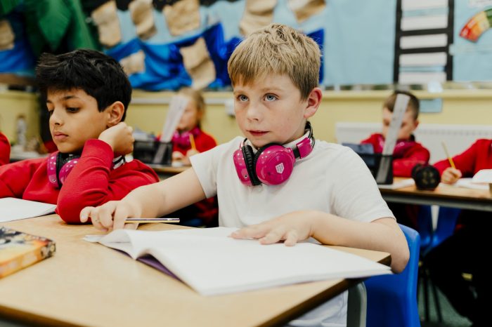 Picture of a boy in class writing while wearing pink Now Press Play headphones.