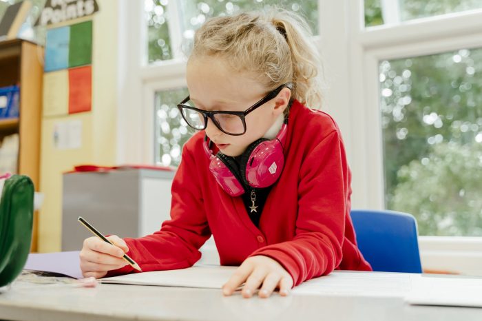 A girl writing in her classroom while wearing Now Press Play headphones