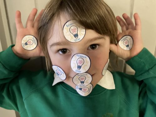 A school child in a green jumper makes a face at the camera. He's covered in Now Press Play Science Star stickers