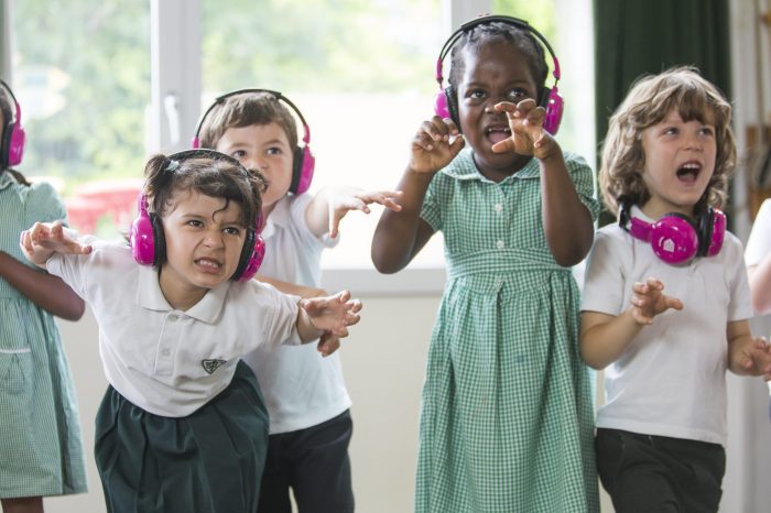 A group of school children act out being dinosaurs during their Now Press Play Experience. They are wearing pink headphones and snarling.