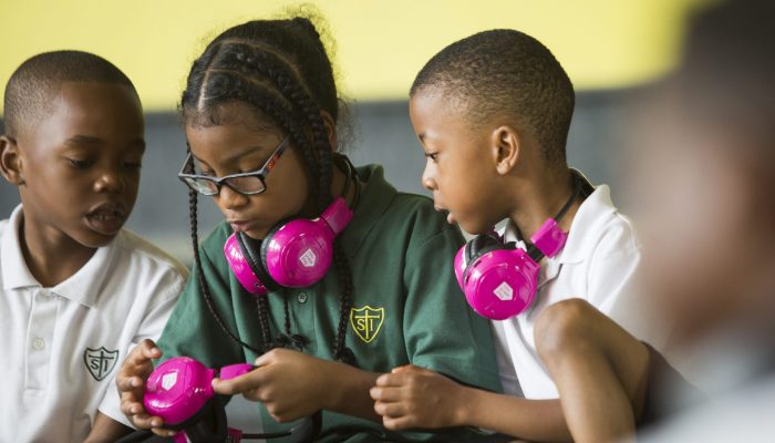 Three children use Now Press Play. Two have pink headphones around their necks, and once is showing another how to adjust the headphones.