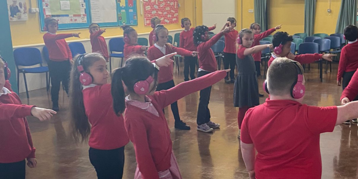 A group of school pupils point as they use Now Press Play. They are in a school hall, wearing a red school uniform, and wearing pink headphones.