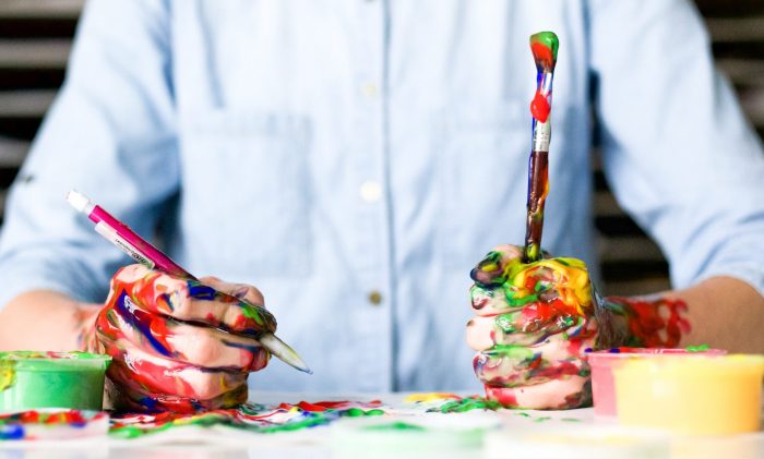 A person holds two paintbrushes, with paint all over their hands.
