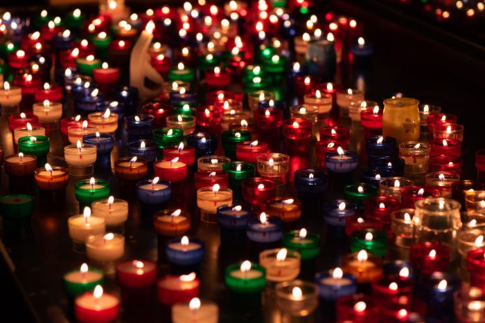 A number of coloured candles lit and standing on the ground at night.