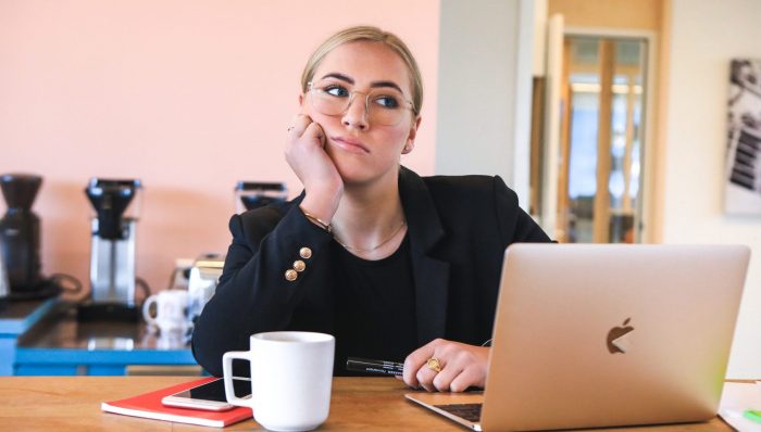 A woman wearing black is sitting at a table, looking fed up. She has a laptop out and a mug of coffee.
